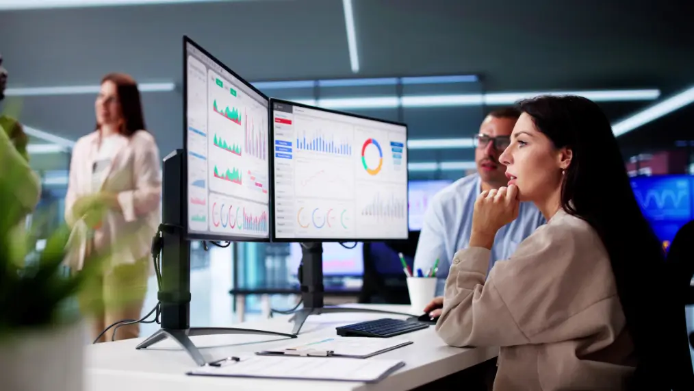 Two analysts reviewing charts and financial dashboards on dual monitors in an office.